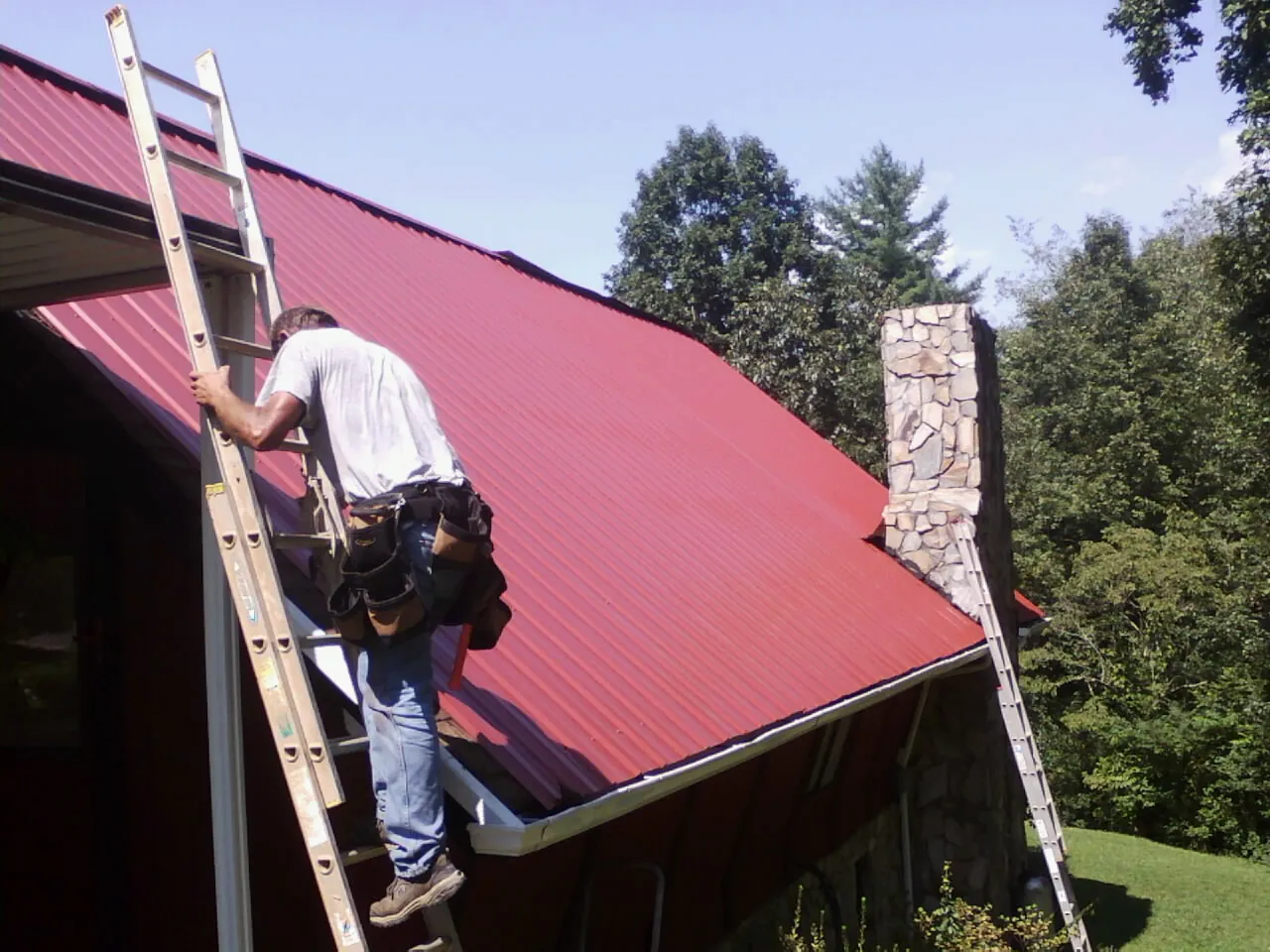 Roofer on a ladder beside a red metal roof and stone chimney — Soffit & Fascia Repair by Roofing Brothers in Hopedale